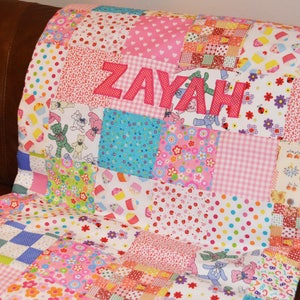 May include: A patchwork quilt with the name "ZAYAH" appliqu&eacute;d in pink letters. The quilt features a variety of colorful fabric squares with patterns including hearts, polka dots, teddy bears, and floral designs. The quilt is resting on a brown surface.