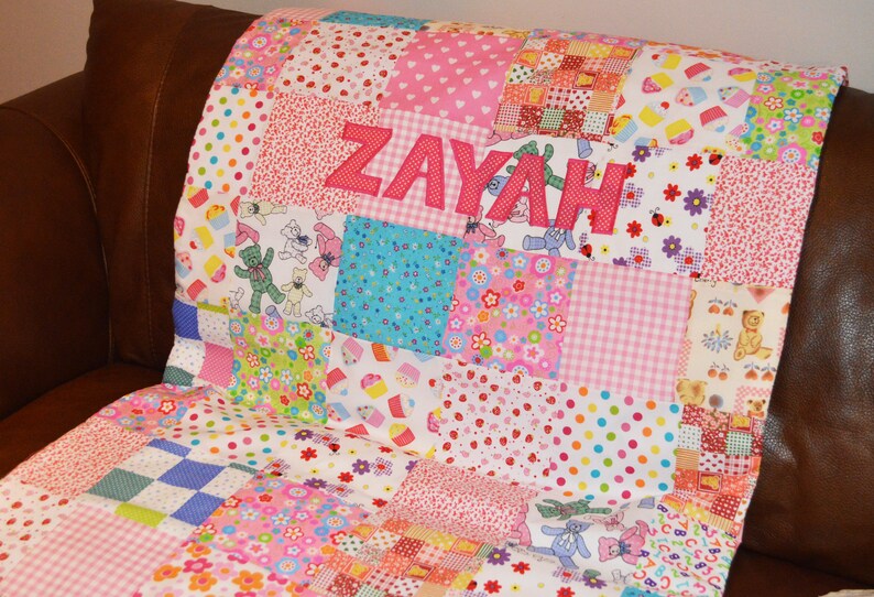 May include: A patchwork quilt with the name "ZAYAH" appliqu&eacute;d in pink letters. The quilt features a variety of colorful fabric squares with patterns including hearts, polka dots, teddy bears, and floral designs. The quilt is resting on a brown surface.
