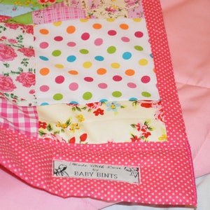 May include: A patchwork quilt with a pink polka dot border and backing. The quilt features various floral and polka dot patterns in pink, white, and other colors. The label reads "Made with Love by BABY BINTS."