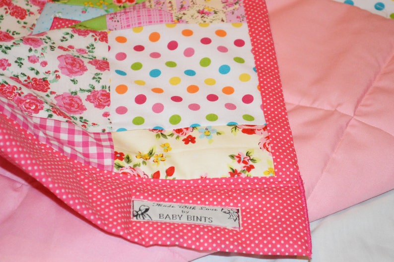 May include: A patchwork quilt with a pink polka dot border and backing. The quilt features various floral and polka dot patterns in pink, white, and other colors. The label reads "Made with Love by BABY BINTS."
