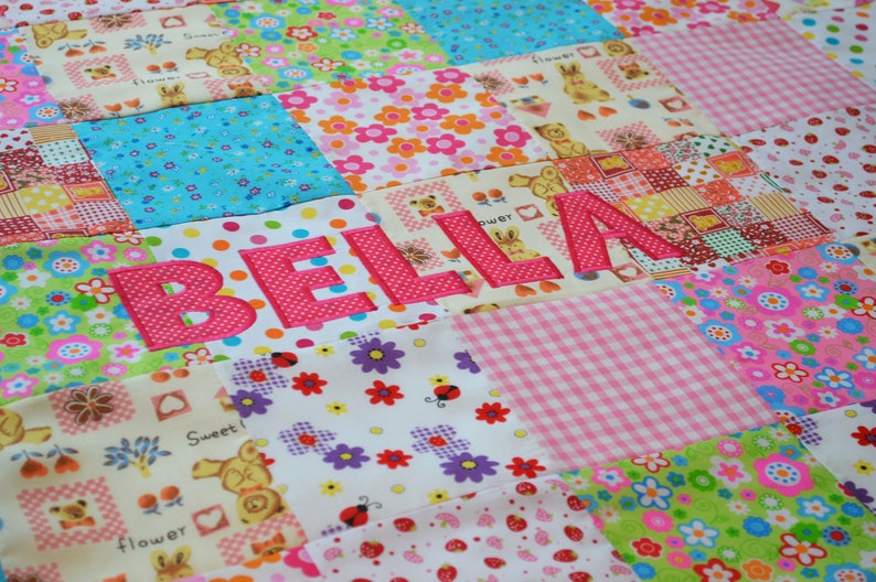 May include: A patchwork quilt with a variety of colorful floral, polka dot, and patterned fabrics. The name "BELLA" is appliqu&eacute;d in pink, adding a personalized touch. The quilt is a mix of pink, blue, green, and yellow squares.