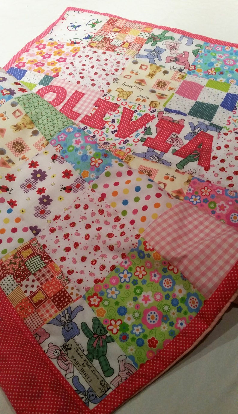 May include: A patchwork quilt featuring a variety of colorful fabrics with floral, polka dot, and teddy bear patterns. The name "OLIVIA" is prominently displayed in large, red, polka-dotted letters. The quilt is trimmed with a red polka dot border.