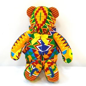 May include: A colourful teddy bear with a yellow bow tie. The bear is made of fabric with a geometric pattern in shades of red, yellow, green, and blue.
