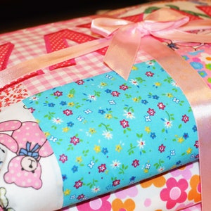 May include: A stack of colorful fabric items tied with a pink satin ribbon. The fabrics feature various patterns, including gingham, floral, and a bunny print. The top layer has the name "Kayla" appliqued in red.