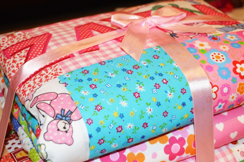 May include: A stack of colorful fabric items tied with a pink satin ribbon. The fabrics feature various patterns, including gingham, floral, and a bunny print. The top layer has the name "Kayla" appliqued in red.