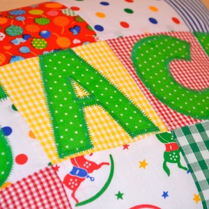 May include: A patchwork pillow with the name "JACKI" appliqued in green polka-dot letters. The pillow features various fabric patterns, including red with balloons, yellow gingham, and red and white gingham.