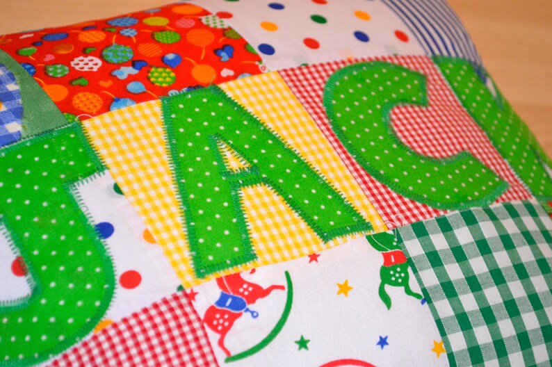 May include: A patchwork pillow with the name "JACKI" appliqued in green polka-dot letters. The pillow features various fabric patterns, including red with balloons, yellow gingham, and red and white gingham.
