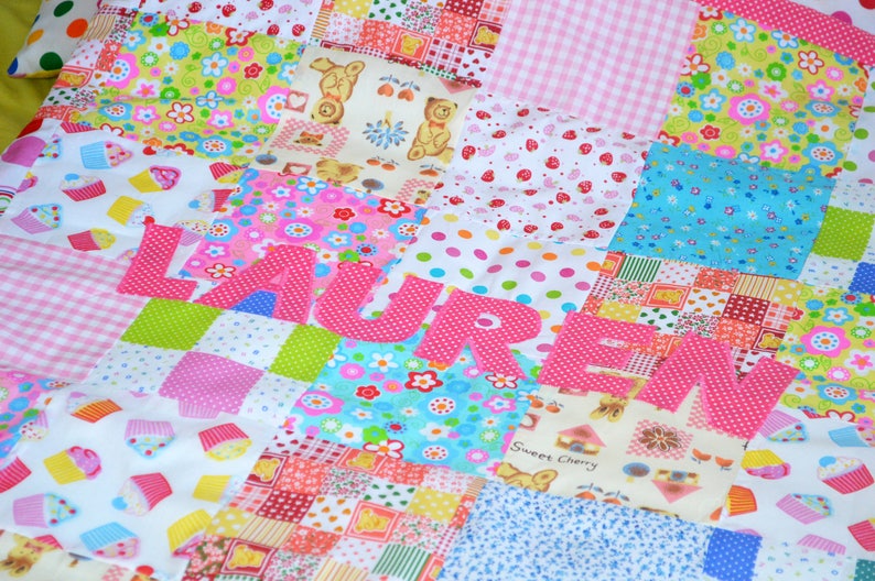 May include: A colorful patchwork quilt with the name "LAUREN" appliqu&eacute;d in pink polka dot fabric. The quilt features various patterned squares with floral, cupcake, and teddy bear designs. The color palette includes pink, blue, green, and white.