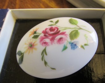 May include: A white oval ceramic trinket box with a floral design. The design features pink, red, yellow, and blue flowers with green leaves.