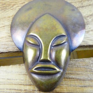 May include: A bronze-colored metal mask with a stylized human face. The mask has a rounded top and a flat, rectangular base.