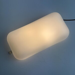 May include: A rectangular wall lamp with a smooth, frosted white glass shade that emits a warm, diffused glow. The fixture features rounded corners and a thin black power cord extending from the side, with a small metallic mounting bracket visible.