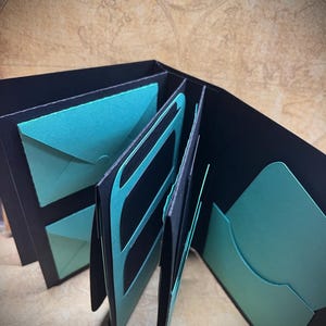 May include: A black leather-bound journal with teal blue paper inserts. The journal has multiple pockets and slots for storing items.