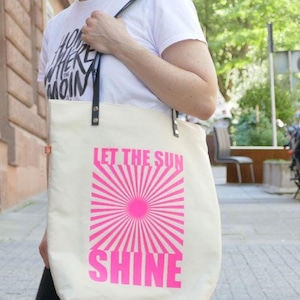 Könnte beinhalten: Eine weiße Canvas-Tragetasche mit schwarzen Ledergriffen und einem rosafarbenen Sonnenstrahlen-Design. Die Tasche trägt den Text "Let the Sun Shine".