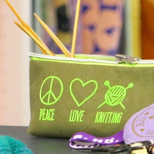 Könnte beinhalten: Eine grüne Canvas-Reißverschlusstasche mit den Worten "Peace, Love, Knitting" und einer Grafik mit Friedenszeichen, Herz und Stricknadeln. Die Tasche ist mit Stricknadeln aus Holz gefüllt.