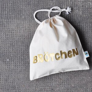 Könnte beinhalten: Eine weiße Baumwolltasche mit Kordelzug, auf der das Wort "Brötchen" in goldenen Buchstaben gedruckt ist. Die Tasche hat ein kleines grünes und blaues Etikett an der Seite.