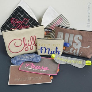 Könnte beinhalten: Ein Set aus sechs braunen Canvas-Reißverschlusstaschen mit verschiedenen gestickten Sprüchen, darunter "Chill Out", "Zeit Für Mich", "Aus Zeit" und "Pause". Die Taschen sind mit goldenem Glitzer, rosa und blauer Stickerei sowie gelben und rosa Borten verziert. Die Taschen sind auf einer braunen Oberfläche angeordnet.