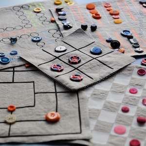 Könnte beinhalten: Fünf verschiedene Stoffquadrate mit verschiedenen darauf gezeichneten Spielen. Jedes Quadrat hat Knöpfe in verschiedenen Farben und Größen, die auf den Spielbrettern platziert sind. Zu den Spielen gehören Tic-Tac-Toe, Dame, ein Labyrinth und ein Punkte-Verbindungsspiel.