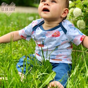 Może przedstawiać: Dziecko w białej koszulce z krótkim rękawem z kwiatowym nadrukiem i niebieskich dżinsach. Koszulka ma niebieską lamówkę wokół dekoltu. Dziecko siedzi na trawie i patrzy w górę.