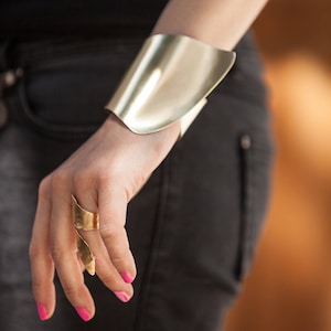 Peut inclure: Un bracelet de manchette en métal doré avec un design incurvé. Le bracelet est porté au poignet d'une personne portant un pantalon noir.