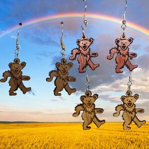 May include: Six pairs of wooden earrings featuring a cartoon bear design. The bears are brown and have a whimsical expression. The earrings are hanging from silver hooks.