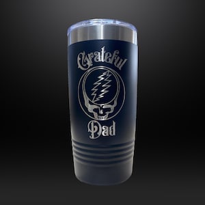 May include: A navy blue 20 oz tumbler with a silver lid. The tumbler features the words "Grateful Dad" in a decorative font, along with a skull and lightning bolt design. The tumbler has horizontal lines near the bottom.