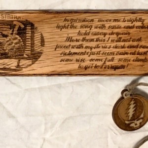 May include: Wooden bookmark with a laser-etched image of two figures and a quote: "Inspiration moves me brightly light the song with gentle cand color hold usery de more than this I will not ack facest with my ste ries berk end omel Halements just seene bazirend bust seme rise some full some limbs to get to Ferique". The bookmark is attached to a brown leather cord with a round charm featuring a lightning bolt design.