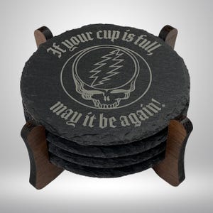 May include: A set of round, dark gray slate coasters with a wooden holder. Each coaster features the text "If your cup is full, may it be again!" around a skull and lightning bolt design.