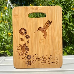 May include: A brown bamboo cutting board with a hummingbird and floral design engraved on it. The board has a handle and the word "Grateful" is engraved on the bottom.
