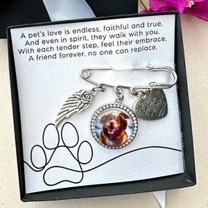 May include: A black box with a white ribbon and a white card inside. The card has a paw print, a silver angel wing charm, a silver heart charm with the text "You'll always be my furry angel", and a silver round charm with a photo of a brown dog. The text on the card reads "A pet's love is endless, faithful and true, And even in spirit, they walk with you. With each tender step, feel their embrace, A friend forever, no one can replace."