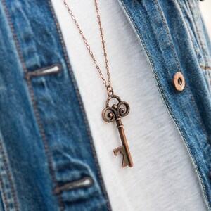 May include: A copper key-shaped bottle opener on a chain necklace. The necklace is worn over a white t-shirt and a blue denim jacket.