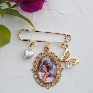 以下が含まれることがあります： 写真チャーム、パールハートチャーム、バタフライチャームが付いたゴールドの安全ピン。写真チャームには、フレーム付きの楕円形の中にカップルが写っています。