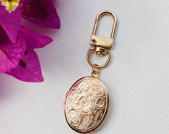 Könnte beinhalten: Ein goldfarbenes ovales Medaillon mit floralem Design, befestigt an einem goldfarbenen Verschluss. Das Medaillon ist an einem kleinen Ring befestigt, der mit dem Verschluss verbunden ist. Der Hintergrund ist eine weiße Oberfläche mit einer lila Blume in der oberen linken Ecke.