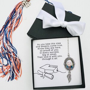 May include: A black box with a white ribbon and a graduation cap illustration. Inside the box is a silver necklace with a photo charm and a feather charm. The necklace is accompanied by a card with the text "As you take this step and dreams come true, Know they are here, so proud of you. In every success, big or small, Their love is with you, through it all."