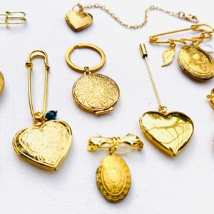 May include: A collection of gold-toned lockets and brooches, including heart-shaped and oval designs. Some pieces feature intricate patterns and are attached to safety pins, chains, and key rings. The jewelry is displayed on a white surface.