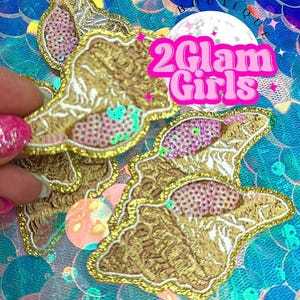 May include: Embroidered patches with gold trim, featuring a detailed design with pink and green accents. The patches are displayed on a shimmering blue sequin background. The text "2Glam Girls" is visible.