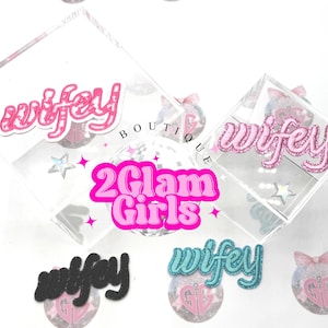 May include: Clear acrylic box with the word "wifey" in pink glitter lettering. The image also features the text "2 Glam Girls" in pink and other decorative elements, including stars and bows.