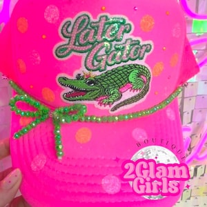 May include: Bright pink trucker hat with the text "Later Gator" and a green alligator wearing a crown. The hat has a green beaded bow and is decorated with orange polka dots and rhinestones. The hat also has the text "2 Glam Girls Boutique".