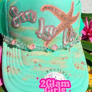 May include: A turquoise trucker hat with a glittery design. The hat features the words "Sea La Vie" in pink and a starfish. A beaded strand is attached to the brim. The bottom of the hat has the text "2 Glam Girls".