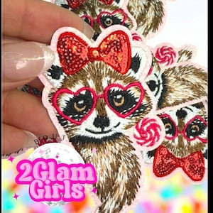 May include: Embroidered patches featuring a raccoon wearing heart-shaped glasses and a bow. The patches are brown, pink, and red, with a lollipop and the text "2 Glam Girls".
