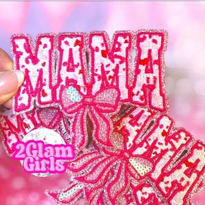 May include: Pink and white sequined "MAMA" patch with a bow design. The patch has a glittery texture and is held by a hand. The text "2 Glam Girls" is also visible.