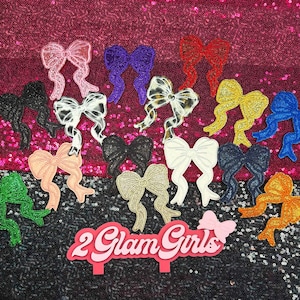 May include: A collection of glittery bow-shaped embellishments in various colors, including pink, purple, red, yellow, and blue. The bows are arranged on a sequined background, with a sign that reads "2 Glam Girls".