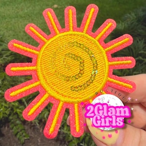 May include: Embroidered sun patch with a yellow center and pink and yellow rays. The center features a gold spiral design. The patch has a stitched border. The text "2 Glam Girls" is visible on a sticker.