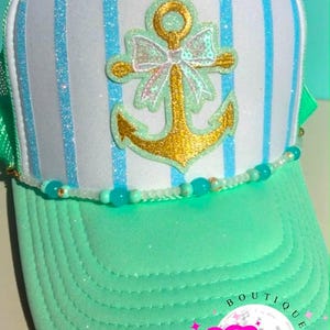 May include: A trucker hat with a white front panel featuring blue glitter stripes. A gold anchor with a bow is centered on the front. The brim is mint green, and a beaded strand accents the brim. The hat has a green mesh back.