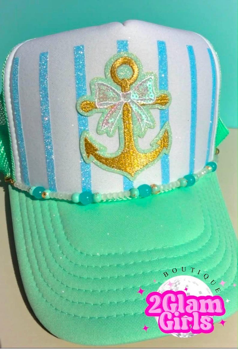 May include: A trucker hat with a white front panel featuring blue glitter stripes. A gold anchor with a bow is centered on the front. The brim is mint green, and a beaded strand accents the brim. The hat has a green mesh back.