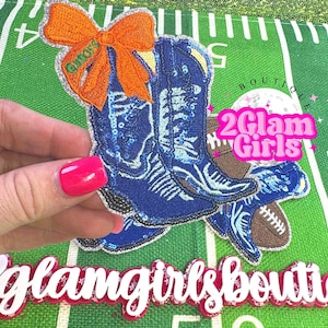 May include: A blue sequined cowboy boot applique with an orange bow and the word "Gators". A brown football is next to the boots. The text "2Glam Girls" is in a pink bubble.