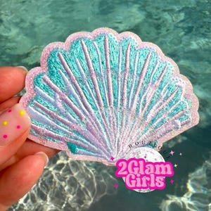 May include: Embroidered seashell patch in shades of teal, pink, and lavender. The patch has a scalloped edge and detailed stitching, with the text "2Glam Girls" in a pink and white design.