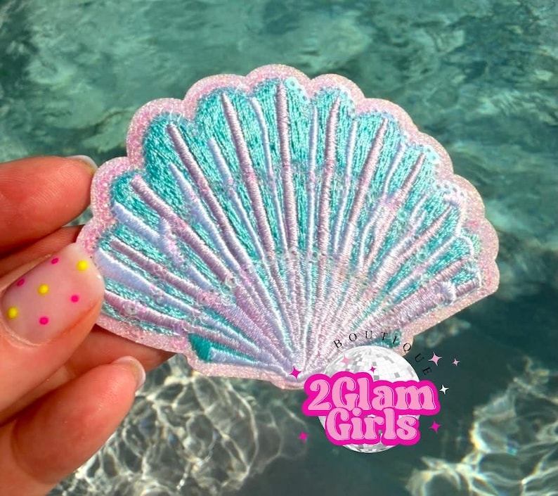 May include: Embroidered seashell patch in teal, pink, and lavender. The patch has a scalloped edge and detailed stitching, with the text "2Glam Girls" in a pink and white design.
