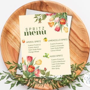 Może przedstawiać: Karta menu z tytułem "SPRITZ menu" i przepisami na koktajle, w tym Aperol, Limoncello i Hugo Spritz. Karta jest ozdobiona akwarelowymi kwiatami i spoczywa na drewnianym talerzu z zielenią.