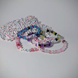May include: A collection of beaded bracelets in various colors, including pink, purple, blue, and green. Each bracelet features letter beads spelling out words. A pile of white letter beads is in the background.