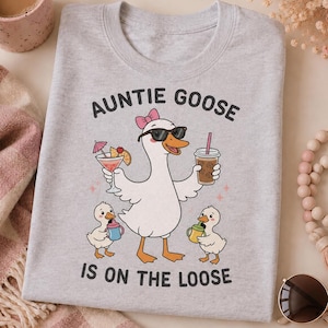 May include: A light gray t-shirt featuring a graphic of a white goose wearing sunglasses and a pink bow, holding a cocktail and a coffee cup. Two small goslings holding sippy cups stand below. The text reads AUNTIE GOOSE IS ON THE LOOSE.